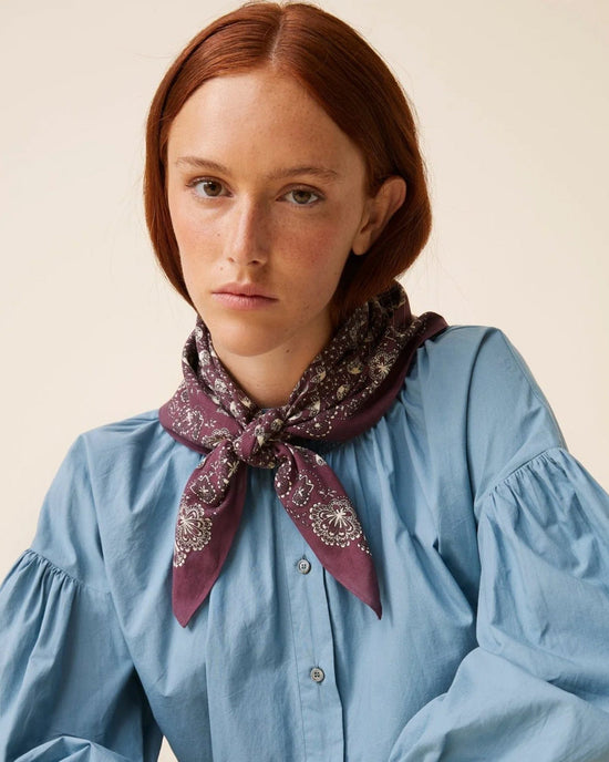 A person with red hair wearing a blue cotton blouse and a Moismont Bandana No 675 in Plum, looking at the camera.