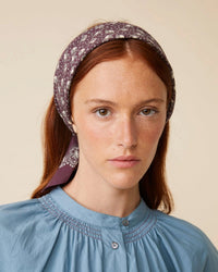 Person with long red hair and a Bandana No 675 in Plum by Moismont, wearing a screen-printed blue top with smocking detail, neutral expression.