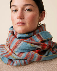 A person is wearing Moismont's Scarf No 699 in Cloud, a multicolored, striped hand-woven wool scarf, along with a beige sweater. They have a neutral expression and are accessorized with hoop earrings against a plain background.