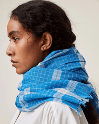 A person with dark hair is shown in profile wearing a Moismont Scarf No 725 in Blue Worker and a white shirt against a plain background.