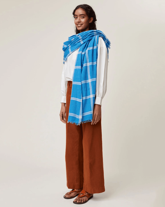 A person wearing a white shirt, brown wide-leg pants, and sandals, with a Moismont Scarf No 725 in Blue Worker draped over their shoulders, stands against a plain background.