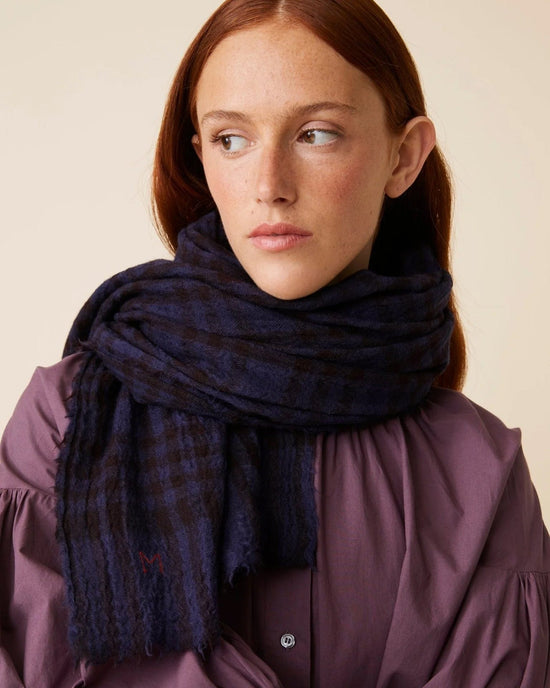 A woman with long red hair wearing a purple top and the exquisite Moismont Scarf No 754 in navy blue, a dark plaid hand-woven wool scarf, looks to the side against a neutral background.