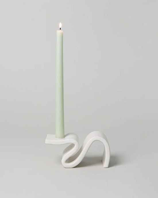 A lit white taper candle is placed in the Wei Candlestick in Cream by SIN, with its wavy, modern design set against a plain background, capturing the essence of contemporary architecture.