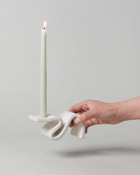 A hand holding the Wei Candlestick in Cream by SIN, featuring a lit, pale green candle.