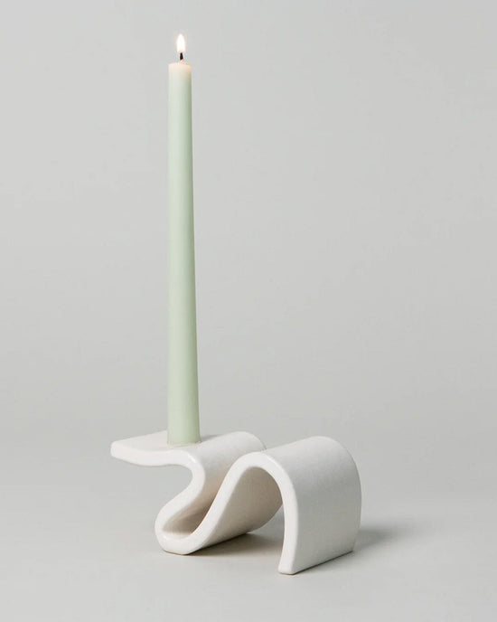 A lit green candle sits elegantly in the Wei Candlestick in Cream by SIN against a plain background, reminiscent of contemporary architecture.