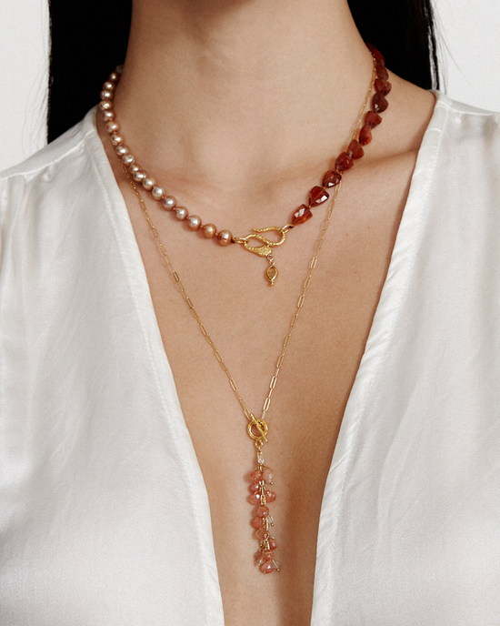 A person wearing a white blouse and two necklaces: a champagne pearl necklace with a gold clasp, and an 18k gold plated sterling silver chain with a pendant featuring small hessonite gemstones, specifically the CL NG-15225LQ in Natural Mix by Chan Luu.