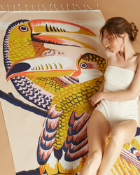 Woman relaxing next to a colorful bird illustration, wrapped in a Inoui Editions Fouta 100 Toucan in Nude 100% Cotton Towel.