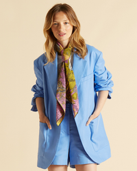 A woman wearing a blue blazer and shorts with a patterned Inoui Editions Square 100 C/S Reverie in Yellow tied around her neck.