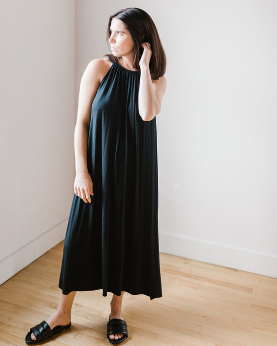 Woman in a Reeba Halter Maxi Dress in Black by Velvet by Graham & Spencer, standing in a room with light walls and wooden floor, looking to the side.