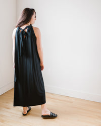 Woman standing in a room wearing a Reeba Halter Maxi Dress in Black with a bow at the back from Velvet by Graham & Spencer.