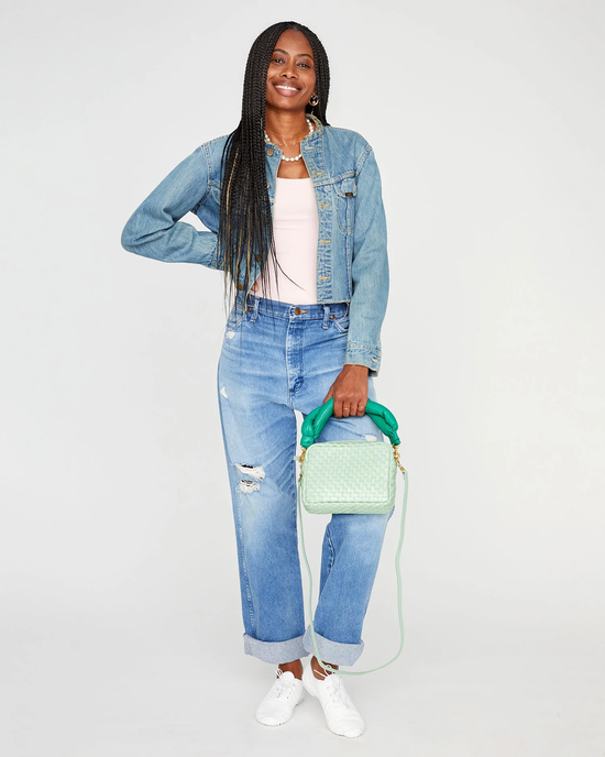Woman in denim jacket and jeans holding a green Clare V. Midi Sac in Mist Woven Checker with a crossbody strap smiles at the camera.