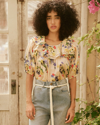 A woman with curly hair standing in front of a door, wearing The Sunrise Top in Bright Grove Floral by the Great and high-rise denim, with a contemplative expression.