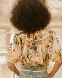 A person with curly hair wearing the Sunrise Top in Bright Grove Floral by the Great, along with high-rise denim jeans viewed from the back.