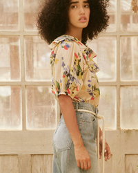 A woman in the Great's Sunrise Top in Bright Grove Floral and high-rise denim jeans standing by a window.