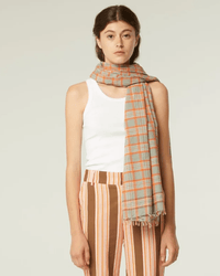 A person wearing a white tank top, striped pants, and the Varied Plaid Cotton Scarf in Cloud from Moismont stands against a plain background.